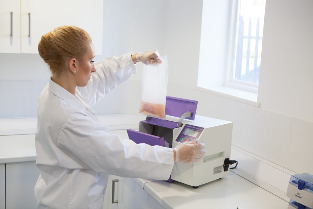 Lab technician using a Stomacher 400 with sample bag, bag rack, and bag opener during sample preparation